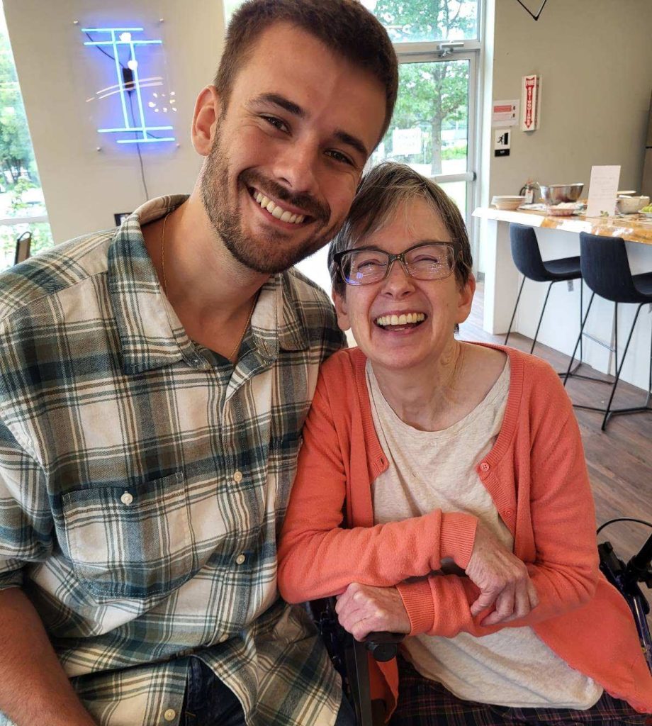 image: smiling young man in plaid shirt sitting close to a smiling woman in orange sweater sitting in a wheelchair